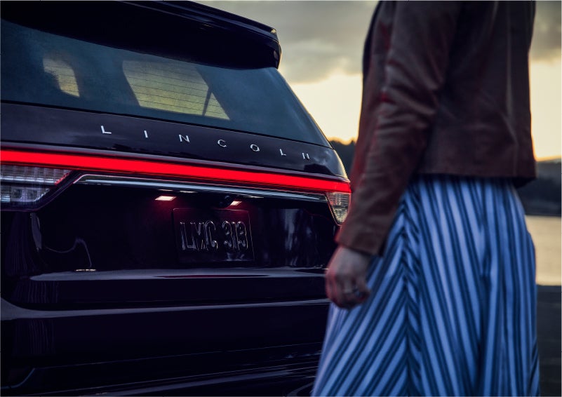A person is shown near the rear of a 2023 Lincoln Aviator® SUV as the Lincoln Embrace illuminates the rear lights | Miller Lincoln in Saint Cloud MN